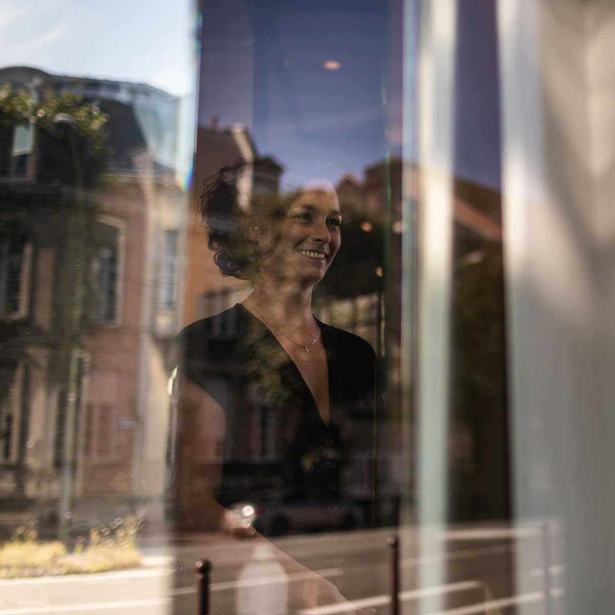 Mariée derrière une vitre qui sourit lors de ses préparatifs, dans le reflet on aperçoit le ciel et des bâtiments