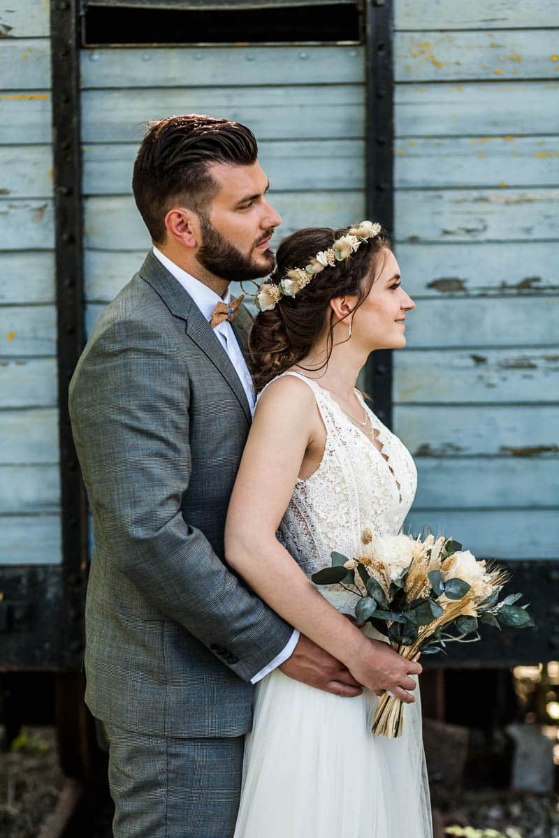 Portrait d’un couple de marié de profil, le marié est derrière la mariée qui tient son bouquet lors d’une séance de couple