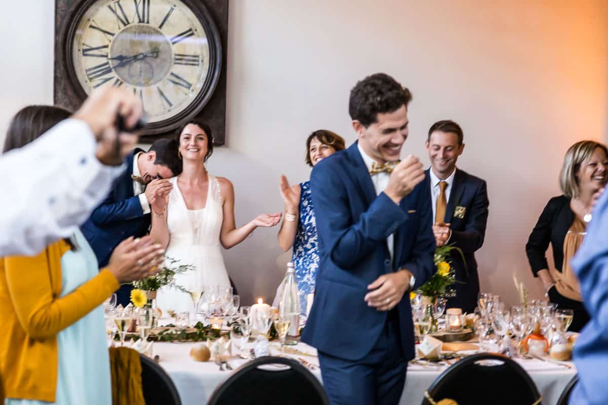 Couple de mariés au second plan et leurs invités ayant un fou rire dans une salle de réception à Lille
