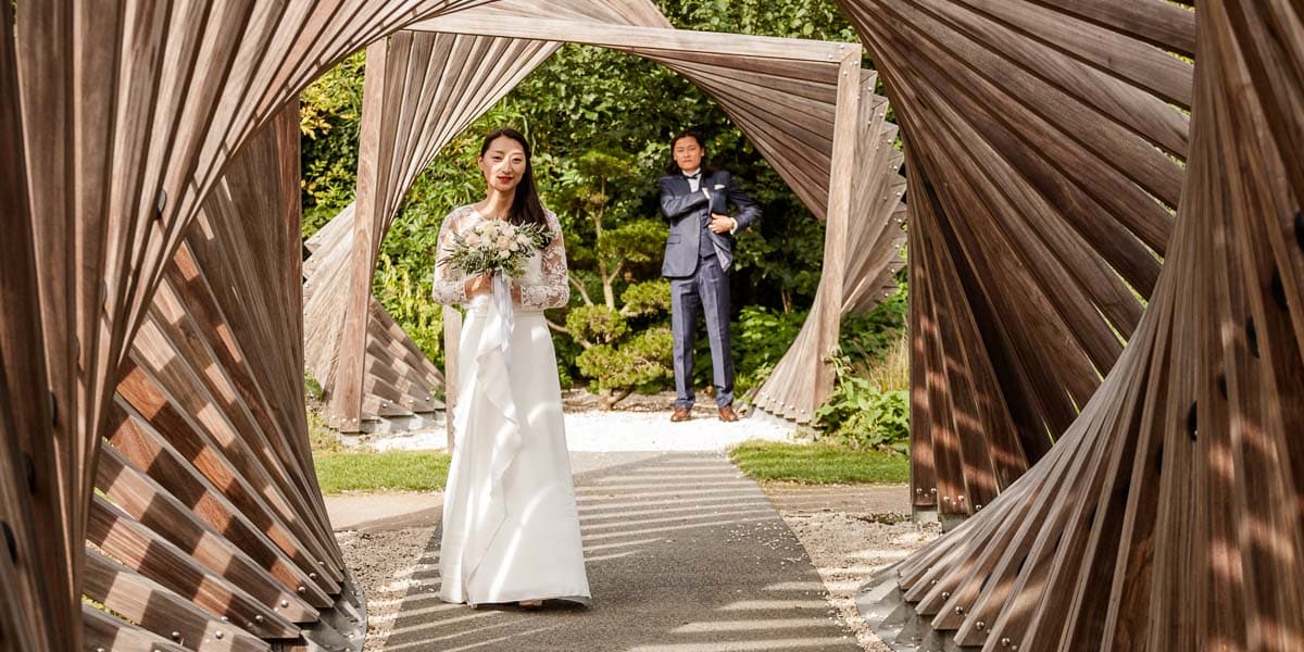 Séance avec un photographe, la mariée est au 1er plan au milieu d’une structure géométrique en bois alors que le marié est au 2nd plan
